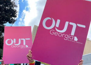 Signs for OUT Georgia Business Alliance written in white on fuchsia held high with sky and tree inthe background.